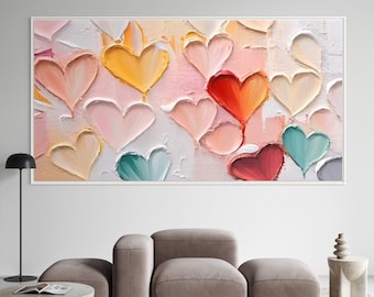 May include: A framed abstract painting featuring textured, colorful hearts in shades of pink, yellow, red, and teal. The artwork has a white border and is displayed on a white wall above a modern living room setup.
