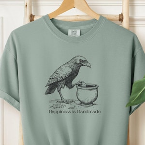 Puede incluir: Una camiseta verde salvia con una ilustración en blanco y negro de un cuervo sosteniendo un caldero. El texto "Happiness is Handmade" está impreso debajo de la ilustración.