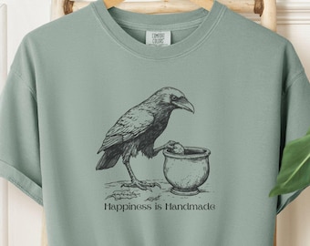May include: A sage green t-shirt with a black and white illustration of a crow holding a cauldron. The text "Happiness is Handmade" is printed below the illustration.