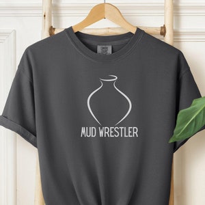 May include: A dark grey t-shirt with a white outline of a vase and the text "MUD WRESTLER" printed on the front.