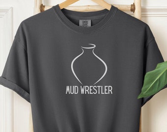 May include: A dark gray t-shirt with a white outline of a vase and the text "MUD WRESTLER" printed on the front.