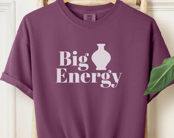 May include: A plum-colored t-shirt with the words "Big Energy" and a vase graphic in white. The shirt is knotted at the waist and hangs on a wooden ladder. A woven basket and a round straw bag are visible in the background.
