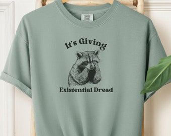 May include: Sage green t-shirt with a graphic of a raccoon and the text "It's Giving Existential Dread." The shirt is hanging on a wooden rack. The t-shirt is knotted at the bottom.
