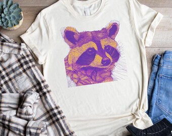May include: A white t-shirt with a purple and yellow illustration of a raccoon's face. The shirt is paired with blue jeans, a plaid shirt, and black ankle boots.