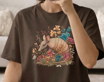 May include: A brown t-shirt featuring a colorful illustration of an armadillo surrounded by flowers. The armadillo has a tan body and is nestled among pink, yellow, and blue flowers. The shirt is a solid brown color.