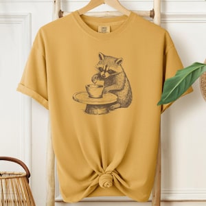 Puede incluir: Una camiseta amarilla mostaza con un gráfico de un mapache usando un torno de alfarería. El mapache es marrón y negro y lleva un delantal blanco.