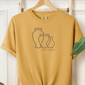 May include: A mustard yellow t-shirt with a graphic of three stylised vases and the text "Pot Dealer."