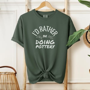May include: A dark green t-shirt with the text "I'd rather be doing pottery" in white lettering.