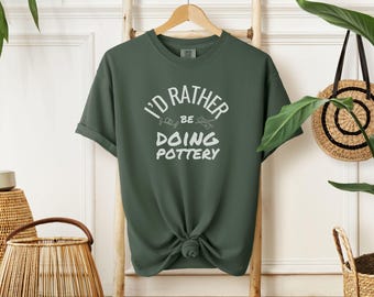 May include: A dark green t-shirt with the text "I'd rather be doing pottery" in white lettering.