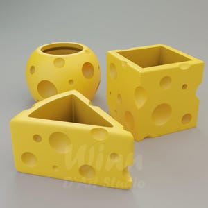 Puede incluir: Tres macetas amarillas con forma de queso. Una es una esfera, otra un cubo y la tercera una cuña triangular. Cada maceta tiene agujeros circulares, que se asemejan al queso suizo. Las macetas están sobre una superficie gris.