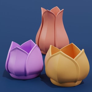 Könnte beinhalten: Drei Keramikvasen in Blumenform in verschiedenen Farben: rosa, lila und gelb. Die Vasen sind auf einem blauen Hintergrund angeordnet.