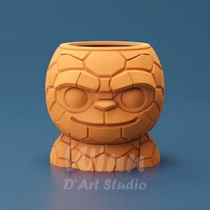 Pode incluir: Jardineira estilizada de cor laranja com design de rosto. A jardineira tem uma aparência texturizada e rochosa e uma expressão sorridente. O texto "D'Art Studio" está na parte inferior. Em um fundo azul.
