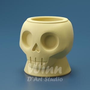 May include: A light yellow skull-shaped planter with large eye sockets and a detailed nose and mouth. The planter has a wide opening at the top for plants. The text "Winn" and "D'Art Studio" are printed on the base.