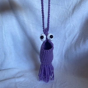 Könnte beinhalten: Lilafarbener gehäkelter Hängebeutel mit weißen Augen und einem Fransenboden.
