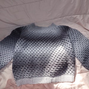 May include: A gray crochet sweater with a crew neck and long sleeves. The sweater has a textured, open-weave design and a ribbed hem and cuffs. The color fades from light to dark gray. The sweater is made of a soft, comfortable material.
