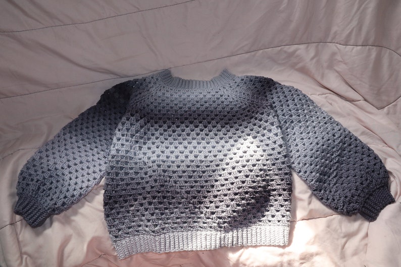 May include: A gray crochet sweater with a crew neck and long sleeves. The sweater has a textured, open-weave design and a ribbed hem and cuffs. The color fades from light to dark gray. The sweater is made of a soft, comfortable material.