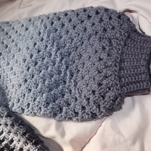 May include: A handmade crochet sweater in shades of gray. The sweater features a textured, open-weave design on the body and sleeves, with a ribbed cuff. The gradient effect transitions from light to dark gray.