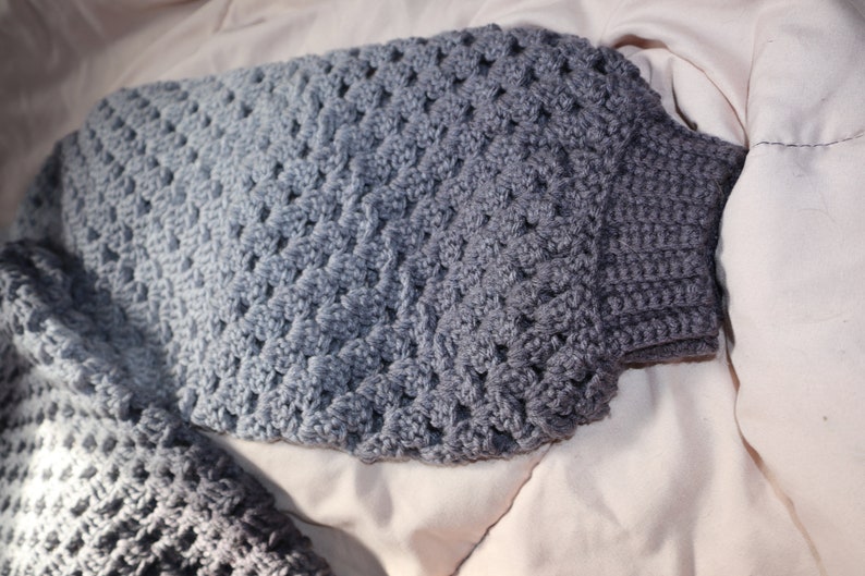May include: A handmade crochet sweater in shades of gray. The sweater features a textured, open-weave design on the body and sleeves, with a ribbed cuff. The gradient effect transitions from light to dark gray.