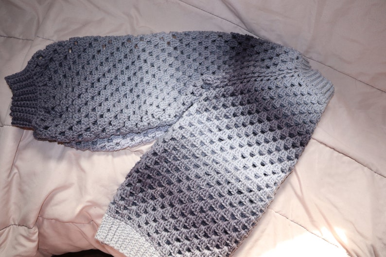 May include: A handmade crochet sweater in shades of gray, transitioning from light to dark. The sweater features a textured, open stitch pattern and ribbed cuffs. The garment is displayed on a light-colored surface.