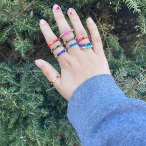 May include: A hand wearing a stack of colorful beaded rings. The rings are made of small, colorful beads strung on a gold wire. The rings are worn on the middle finger of the hand.