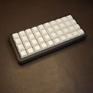 Puede incluir: Un teclado mecánico negro compacto con teclas blancas. Las teclas están dispuestas en una cuadrícula, y el teclado tiene forma rectangular. Las teclas son cuadradas y están espaciadas uniformemente.