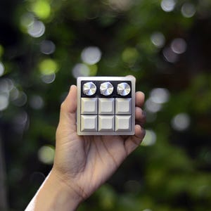 Puede incluir: Un pequeño controlador de música plateado y negro con seis botones y tres perillas. El controlador está siendo sostenido en una mano.