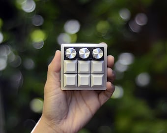 Puede incluir: Un pequeño controlador de música plateado y negro con seis botones y tres perillas. El controlador está siendo sostenido en una mano.