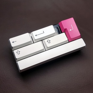 May include: A small, white keyboard with five keys, including arrow keys and a pink key with a right arrow symbol. The keyboard is made of aluminium and has a white finish.