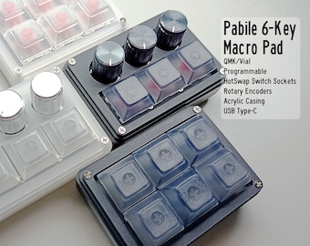 Puede incluir: Un pad de macro negro de 6 teclas con teclas transparentes, dos codificadores rotatorios y el texto "Pabile 6-Key Macro Pad" en blanco. El texto debajo del nombre del producto dice "QMK/Vial Programmable HotSwap Switch Sockets Rotary Encoders Acrylic Casing USB Type-C".