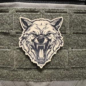 Pode incluir: Um remendo marrom com um gr&aacute;fico de cabe&ccedil;a de lobo em preto e branco. O remendo est&aacute; preso a uma bolsa t&aacute;tica verde.