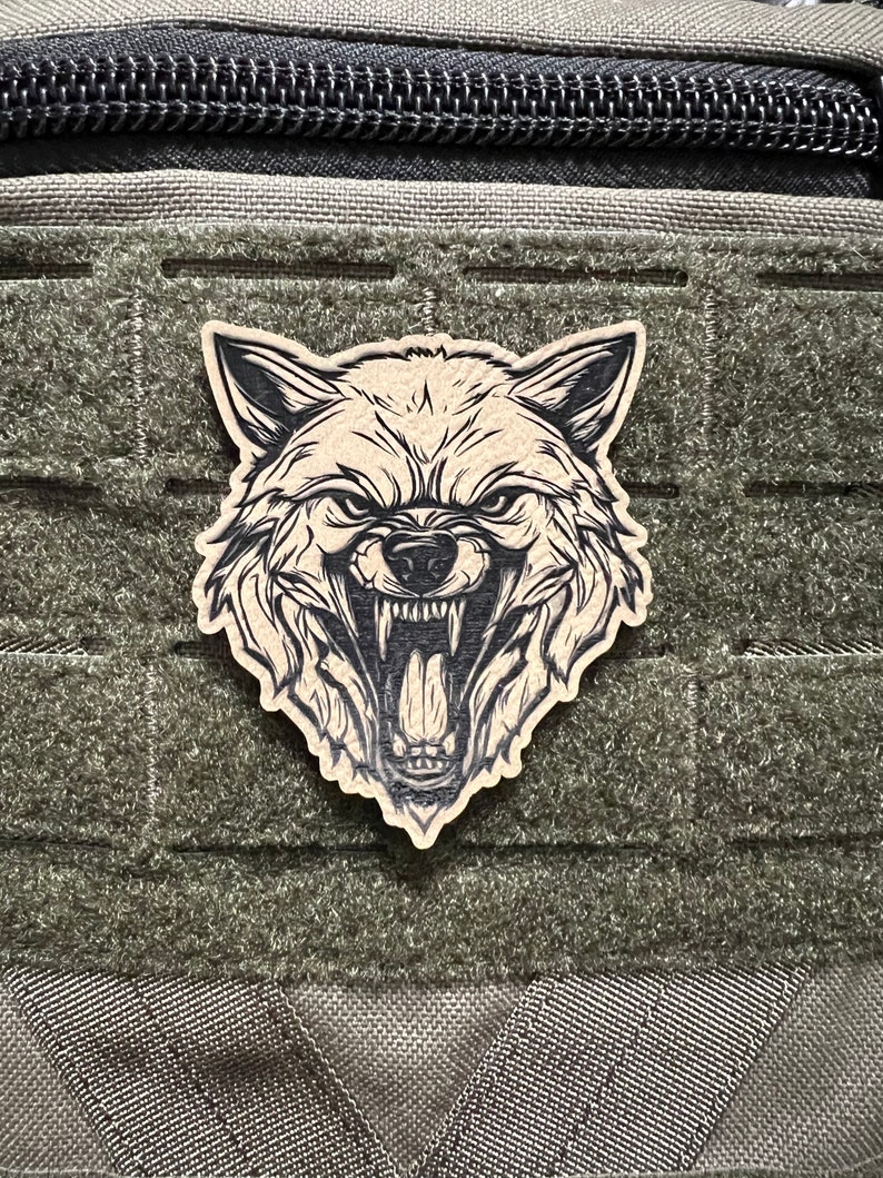 Pode incluir: Um remendo marrom com um gr&aacute;fico de cabe&ccedil;a de lobo em preto e branco. O remendo est&aacute; preso a uma bolsa t&aacute;tica verde.