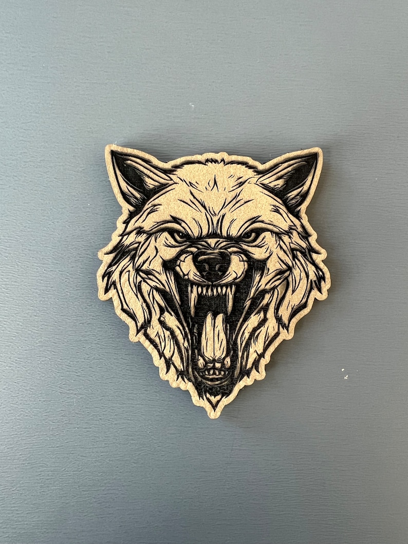 Pode incluir: Um recorte de madeira de uma cabe&ccedil;a de lobo com uma express&atilde;o de rosnado. A pelagem do lobo &eacute; detalhada com linhas pretas e o fundo &eacute; de madeira marrom claro.