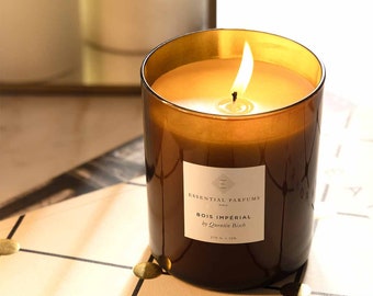 May include: A brown glass candle with a white label that reads "Essential Parfums Paris Bois Imperial by Quentin Bisch 270 g - 12h". The candle is lit and the flame is visible.