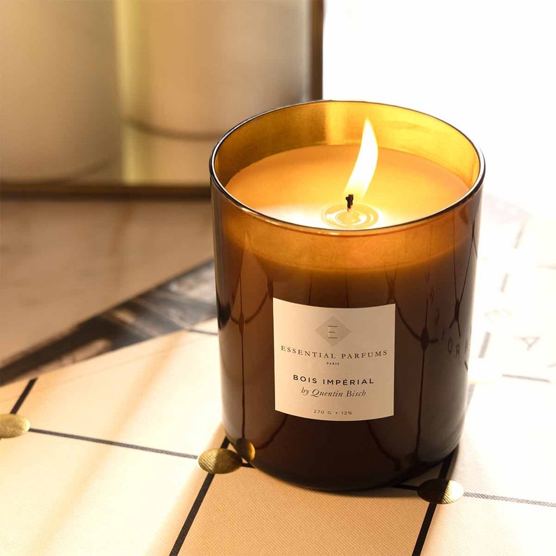 May include: A brown glass candle with a white label that reads "Essential Parfums Paris Bois Imperial by Quentin Bisch 270 g - 12h". The candle is lit and the flame is visible.