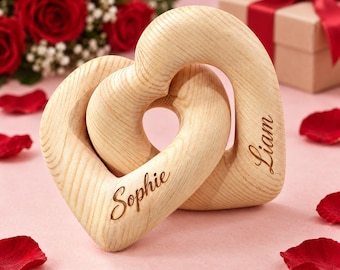 May include: Two interlocking wooden hearts with a light wood grain finish. The left heart is engraved with the name Sophie in a cursive script, and the right heart is engraved with the name Liam in a matching cursive script, forming a single unit.