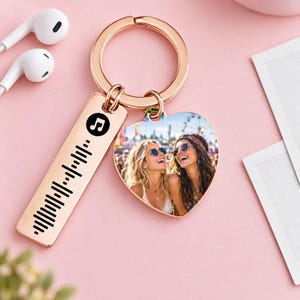 Op de afbeelding: Een roségouden sleutelhanger met een rechthoekig metalen label met een zwart muzieknoot-icoon en een verticale geluidsgolfgrafiek, bevestigd aan een hartvormige bedel met een kleurenfoto van twee lachende mensen met een zonnebril.