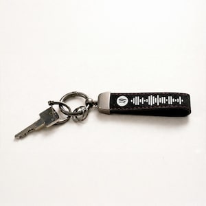Op de afbeelding: Een zwart leren sleutelhanger met een Spotify-code ontwerp en een zilveren sleutel bevestigd. De sleutelhanger heeft een metalen ring en een zwarte band met een witte Spotify-code. De sleutel is zilver.
