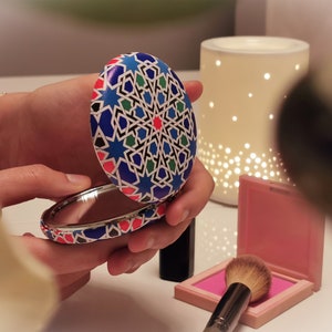 May include: A round, blue, white, red, and green compact mirror with a geometric pattern. The mirror is open and the reflection is visible.
