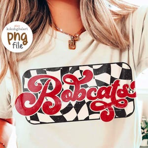 May include: A cream-coloured t-shirt featuring a red and black chequered design with the word "Bobcats" in red script. The person is wearing matching chequered shorts. The image includes a PNG file logo.
