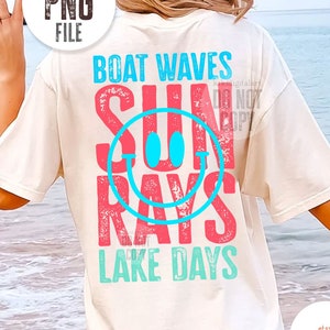 Op de afbeelding: Wit T-shirt met een distressed grafisch ontwerp met de tekst "Boat Waves Sun Rays Lake Days" in roze en blauwe letters. De grafiek bevat een blauwe smiley.