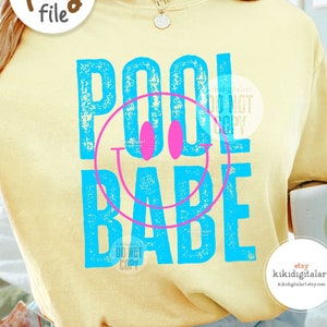 Könnte beinhalten: Ein hellgelbes Sweatshirt mit dem Schriftzug "POOL BABE" in Blau und Pink, verziert mit einem Smiley-Gesicht. Das Sweatshirt wird mit hellblauen Shorts getragen. Eine Kaffeetasse mit einem Smiley ist ebenfalls zu sehen.