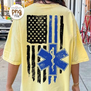 Puede incluir: Camiseta amarilla clara con un diseño de bandera estadounidense desgastada con rayas negras y una franja azul. Un emblema azul de la Estrella de la Vida está centrado debajo. Mangas cortas.