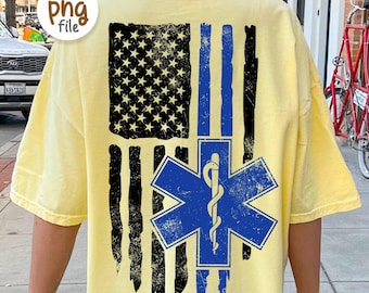 May include: A light yellow t-shirt featuring a distressed American flag design with black stripes and a blue stripe. A blue Star of Life emblem is centered below the flag. The shirt has short sleeves.