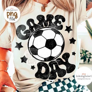 May include: Cream-colored t-shirt with a black and white soccer ball graphic and the words "GAME DAY". The design includes black stars. The shirt is a casual style.