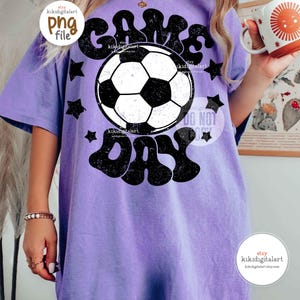 May include: A light purple t-shirt with a black and white soccer ball graphic and the words "GAME DAY". The shirt has a vintage, distressed look with black star accents.