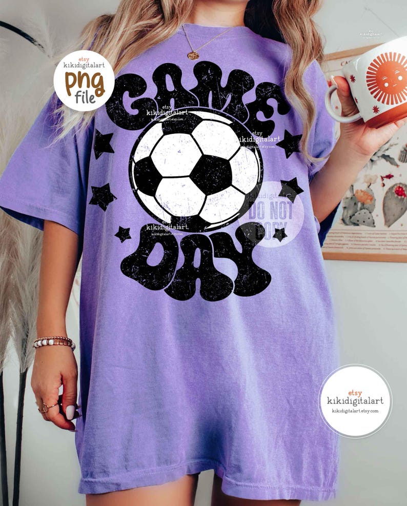 May include: A light purple t-shirt with a black and white soccer ball graphic and the words "GAME DAY". The shirt has a vintage, distressed look with black star accents.