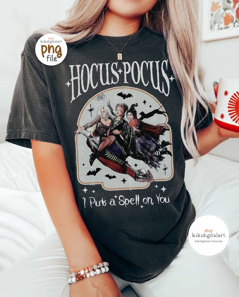 May include: Dark gray t-shirt featuring the text "Hocus Pocus" above a graphic of three witches riding a broom. The phrase "I Put a Spell on You" is printed below the graphic. The shirt has a vintage, distressed look.