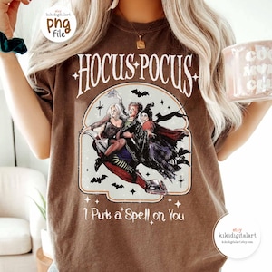 May include: Brown t-shirt featuring the text "Hocus Pocus" with an illustration of three witches riding a broom. The phrase "I Put a Spell on You" is printed below the illustration. The shirt has a vintage style.