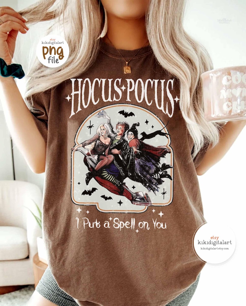May include: Brown t-shirt featuring the text "Hocus Pocus" with an illustration of three witches riding a broom. The phrase "I Put a Spell on You" is printed below the illustration. The shirt has a vintage style.