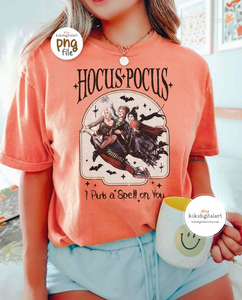 May include: Orange short-sleeved t-shirt featuring the text "Hocus Pocus" above an illustration of three witches riding a broom. The phrase "I Put a Spell on You" is below the illustration. A white mug with a smiley face is held in the hand.